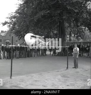 1950er, historisch, beobachtet von einer Reihe von Schuljungen, die draußen auf einem Schulspielplatz standen, ein Teenager, der den Hochsprung machte, über die Bar sprang, kurz bevor er im Sandkasten landete, England, Großbritannien. Stockfoto