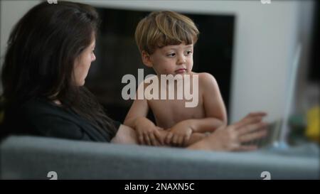 Ein kleiner Junge, der die Aufmerksamkeit der Mutter möchte, während er zu Hause vor einem Laptop arbeitet. Eltern-Multitasking mit Kleinkindern. Erziehungs-Lebensstil-Betrug Stockfoto