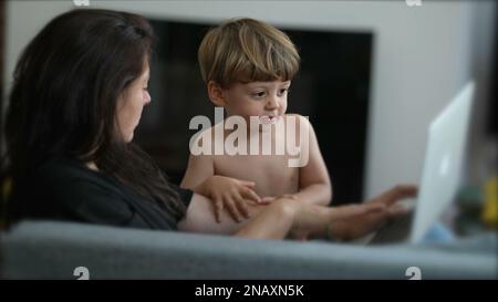 Ein kleiner Junge, der die Aufmerksamkeit der Mutter möchte, während er zu Hause vor einem Laptop arbeitet. Eltern-Multitasking mit Kleinkindern. Erziehungs-Lebensstil-Betrug Stockfoto