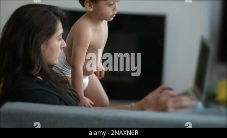 Ein kleiner Junge, der die Aufmerksamkeit der Mutter möchte, während er zu Hause vor einem Laptop arbeitet. Eltern-Multitasking mit Kleinkindern. Erziehungs-Lebensstil-Betrug Stockfoto