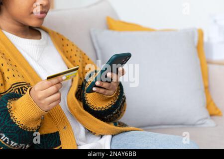 Frau kauft online mit Smartphone und bezahlt mit goldener Kreditkarte. afroamerikanisches Mädchen, das im Internet kauft, Kreditkartendetails eingeben. E-Commerce-Lieferservice für Online-Shopping Stockfoto