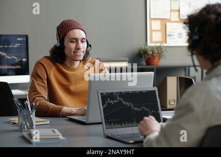 Ein junger, lächelnder männlicher Telefonist in Headset- und Freizeitkleidung, der Online-Kunden berät, während er am Arbeitsplatz vor dem Laptop sitzt Stockfoto