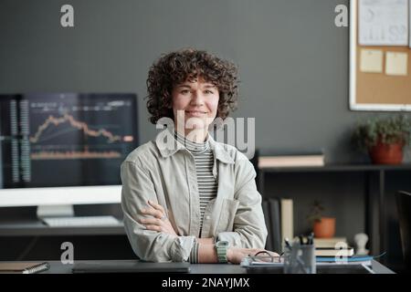 Junge, erfolgreiche Finanzanalytikerin, die am Arbeitsplatz im Büro sitzt und vor die Kamera schaut, um die Grafikdaten auf dem Computerbildschirm abzugleichen Stockfoto