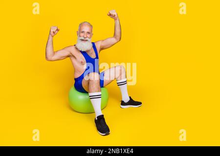 Langes Foto des alten lustigen Großvaters mit Bart Sitzkleidung blaue Sportbekleidung, auf dem die Hände hoch sind starke Muskeln isoliert auf gelber Farbe Stockfoto