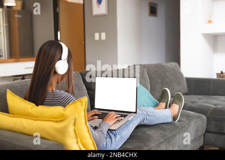 Birassisches Teenager-Mädchen, das Kopfhörer trägt und ein Notebook mit Kopierbereich benutzt. Das Konzept „Zeit zu Hause verbringen“. Stockfoto