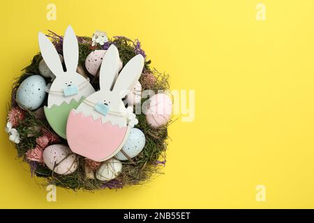 Holzhasen mit Schutzmasken, lackierten Eiern, Osterkranz und Platz für Text auf gelbem Hintergrund, Draufsicht. Feiertag während der COVID-19-Pandemie Stockfoto