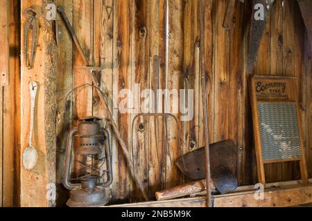 Antike Öllaterne, Heugabel, Schere, Axt, Sägen, Waschbrett und andere Handwerkzeuge hängen an der senkrechten Holzplankenwand in der alten Scheune. Stockfoto