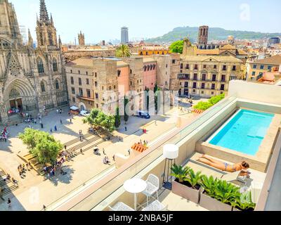 Eine junge Frau sonnt sich neben einem Swimmingpool in einem Luxushotel auf dem Dach mit Blick auf die Kathedrale von Barcelona in Barcelona, Spanien. Stockfoto