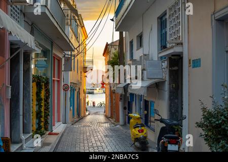 Boote im Hafen bei Sonnenuntergang, von einer engen Gasse mit Geschäften und Häusern auf der griechischen Insel Ägina, Griechenland, eine der Saronischen Inseln aus gesehen Stockfoto