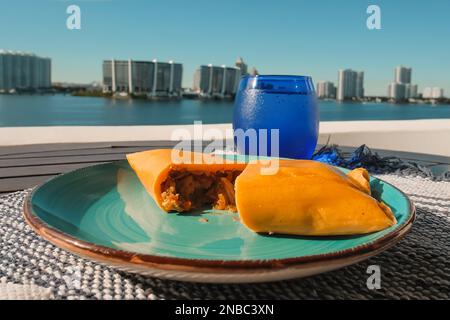 Hallaca oder Tamale über einem rustikalen Tisch und einem blauen Gericht mit einem Getränk, mexikanische und venezolanische traditionelle Speisen mit einem Hintergrund aus Miami, Florida Stockfoto
