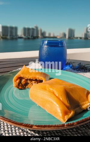 Hallaca oder Tamale über einem rustikalen Tisch und einem blauen Gericht mit einem Getränk, mexikanische und venezolanische traditionelle Speisen mit einem Hintergrund aus Miami, Florida Stockfoto