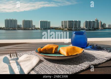 Hallaca oder Tamale über einem rustikalen Tisch und einem blauen Gericht mit einem Getränk, mexikanische und venezolanische traditionelle Speisen mit einem Hintergrund aus Miami, Florida Stockfoto