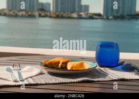 Hallaca oder Tamale über einem rustikalen Tisch und einem blauen Gericht mit einem Getränk, mexikanische und venezolanische traditionelle Speisen mit einem Hintergrund aus Miami, Florida Stockfoto