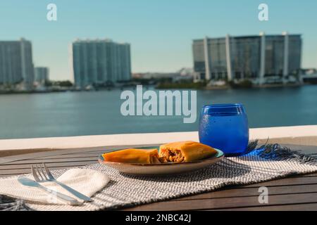 Hallaca oder Tamale über einem rustikalen Tisch und einem blauen Gericht mit einem Getränk, mexikanische und venezolanische traditionelle Speisen mit einem Hintergrund aus Miami, Florida Stockfoto