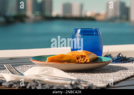 Hallaca oder Tamale über einem rustikalen Tisch und einem blauen Gericht mit einem Getränk, mexikanische und venezolanische traditionelle Speisen mit einem Hintergrund aus Miami, Florida Stockfoto