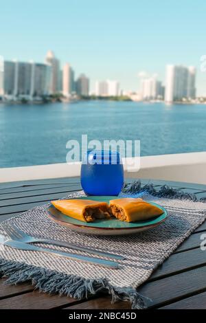 Hallaca oder Tamale über einem rustikalen Tisch und einem blauen Gericht mit einem Getränk, mexikanische und venezolanische traditionelle Speisen mit einem Hintergrund aus Miami, Florida Stockfoto