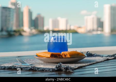 Hallaca oder Tamale über einem rustikalen Tisch und einem blauen Gericht mit einem Getränk, mexikanische und venezolanische traditionelle Speisen mit einem Hintergrund aus Miami, Florida Stockfoto