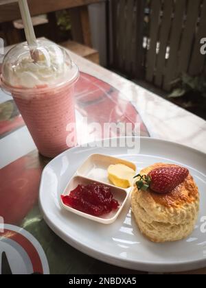Scones auf einem weißen Teller mit Erdbeermarmelade und eine Tasse Erdbeermilchshakes im Hintergrund Stockfoto