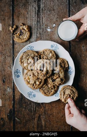 Ein Teller mit Schokoladenkeksen, ein Glas Milch, zwei Hände, die nach Keksen und Milch greifen Stockfoto