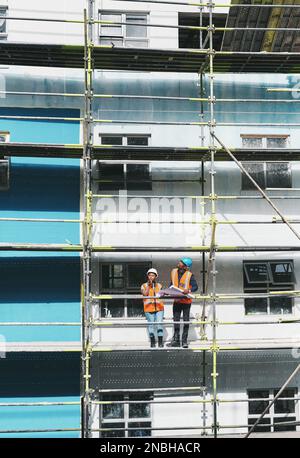 Sicherstellen, dass der Build termingerecht, budgetgerecht und im Rahmen des Umfangs erfolgt. Ein junger Mann und eine junge Frau, die Baupläne auf einer Baustelle durchgehen. Stockfoto