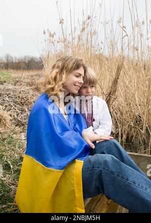 Ein kleiner Junge und eine Frau, eingewickelt in die ukrainische Flagge, umarmen sich gegenseitig, auf der Suche nach Unterstützung. Familie, Flüchtlinge, Einheit, Unterstützung, Patriotismus, Nostalgie. Sto Stockfoto