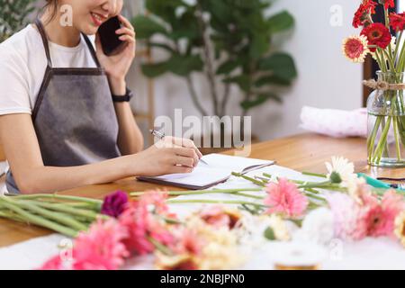 Floristen-Konzept, weibliche Floristin im Gespräch mit dem Kunden am Telefon und Notizen im Notebook im Geschäft. Stockfoto