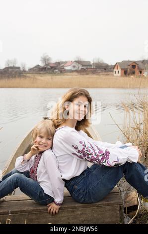 Glücklicher kleiner Junge und junge wunderschöne Frau in bestickter Nationalkleidung sitzen nebeneinander am Ufer des Sees. Familie, Flüchtlinge, Einheit, Unterstützung, Stockfoto