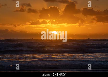 Ein dramatischer orangefarbener Sonnenuntergang, der hinter einer Wolkenschicht aus dem Meer hinausblickt und ein warmes Licht über die unruhigen Wellen des Ozeans wirft. Stockfoto