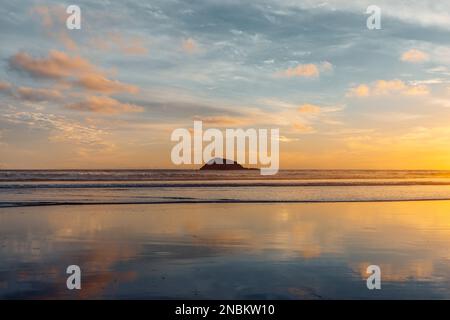 Sonnenuntergangsreflexionen an der Maukatia Bay – Neuseeland mit einer kleinen Oaia-Insel in der Ferne, die sich aus dem Ozean erhebt. Stockfoto