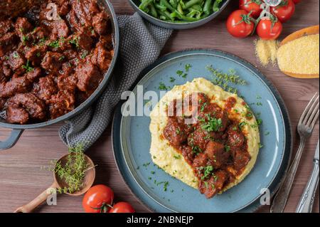 Italienischer Schweineragout mit cremigen Polenta und grünen Bohnen auf Holztisch zum Abendessen oder Mittagessen. Flach verlegt Stockfoto