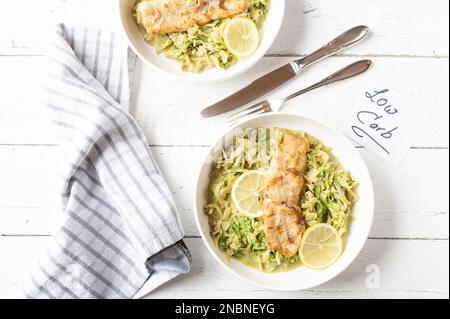 Kohlenhydratarmes Abendessen oder Mittagessen mit gebratenem Fischfilet in der Pfanne und cremigen Zucchini-Nudeln. Serviert auf Tellern auf weißem Hintergrund mit Besteck Stockfoto