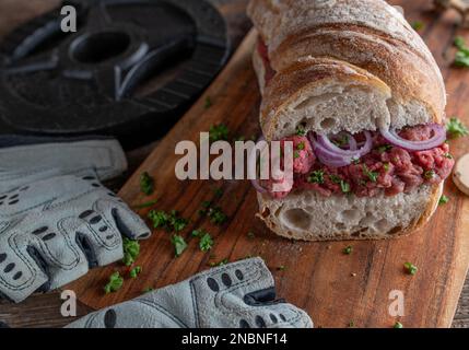 Rohes Rindfleisch-Sandwich auf rustikalem Baguette-Brot mit roten Zwiebeln und Petersilie auf einem Holztisch Stockfoto