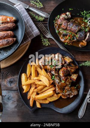 Gebratenes Würstchen mit gebratener deutscher Bratwurst, Zwiebelpilzsauce und hausgemachten pommes frites auf einem Tisch mit Tellern und Pfanne. Flach verlegt Stockfoto