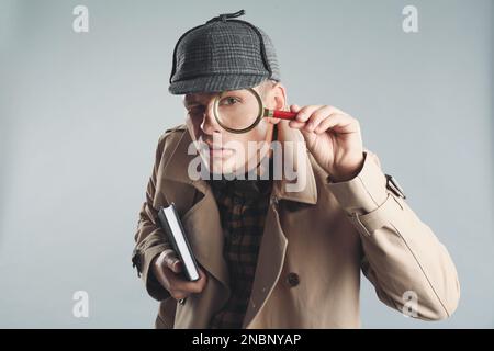 Männlicher Detective, der durch eine Lupe auf grauem Hintergrund schaut Stockfoto