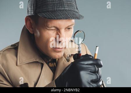 Männlicher Detective, der Zigarettenstummel mit Lupe auf grauem Hintergrund untersucht Stockfoto