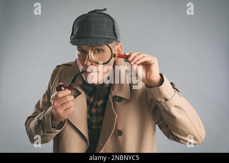 Männlicher Detektiv mit Rauchrohr aus Lupe auf grauem Hintergrund Stockfoto