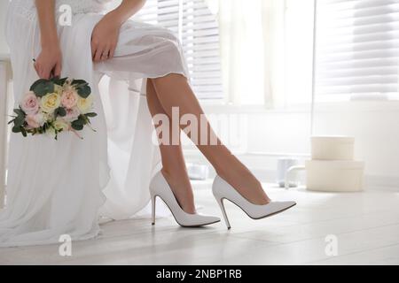 Junge Braut mit Blumenstrauß, die drinnen Hochzeitsschuhe trägt Stockfoto