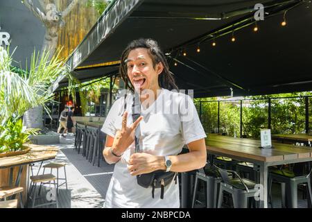 Junger Latino-Mann in einem weißen T-Shirt und Dreadlocks, der vor einem Restaurant steht und auf die Kamera schaut und mit der Hand ein V-Schild macht Stockfoto