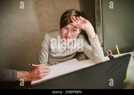 Der Junge schaut zu seinem Vater auf, der ihm bei den Hausaufgaben hilft. Der Schüler beantwortet die Frage des Lehrers, während er online auf einem Laptop lernt. Kinderhand Stockfoto