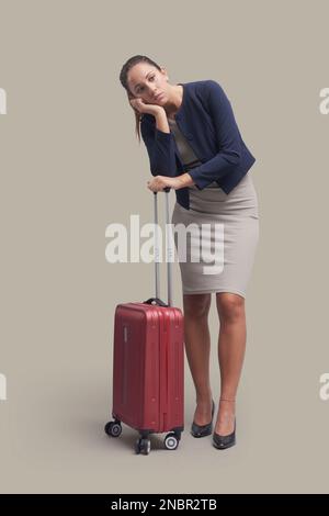 Müde, enttäuschte Geschäftsfrau, die am Bahnhof wartete, lehnt sich an ihren Trolley-Sack-Griff Stockfoto