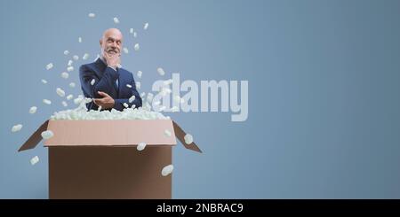 Selbstbewusster lächelnder Geschäftsmann, der mit der Hand am Kinn denkt und aus einer Lieferbox, Geschäftslösungen und einem Kreativitätskonzept herauskommt Stockfoto