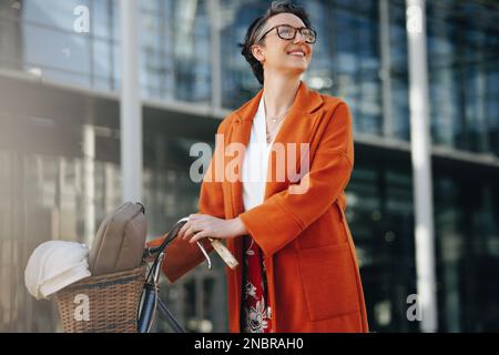 Glückliche Geschäftsfrau, die mit Bedacht wegsieht, während sie ihr Fahrrad in der Stadt schiebt. Lächelnde, reife Geschäftsfrau, die mit einem Fahrrad zur Arbeit pendelt Stockfoto