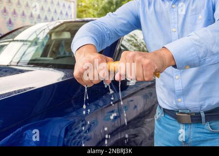 Ein Mann dreht einen Lappen auf, auf einem Hintergrund ein Auto. Ein Mann, der das Auto mit einem Mikrofasertuch reinigt. Der Mensch hält die Mikrofaser in der Hand Stockfoto