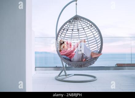Schöne Frau mittleren Alters, barfuß, die in einem bequemen Hängesessel auf der offenen Terrasse liegt und die Sonnenstrahlen des Sonnenuntergangs genießt. Moderner Lebensstil zu Hause Stockfoto
