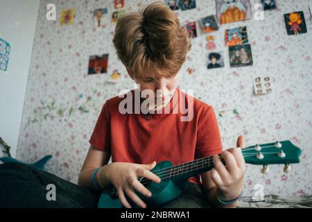 Ein Mädchen im Teenageralter, das auf dem Bett sitzt, lernt Ukulele zu spielen. Stockfoto