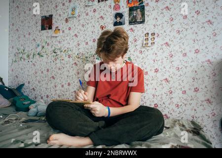 Ein Teenager, das auf einem Bett sitzt, zeichnet ein Skizzenbuch. Stockfoto