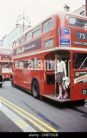 London 1972. Blick auf die Oxford Street mit den typischen roten Doppeldeckerbussen. Eine junge Frau wird auf dem Bahnsteig gesehen, bereit zum Abheben. Sie trägt ein typisches 1970er-Outfit, einen Pullover, einen kurzen rosa gemusterten Rock und dazu passende pinkfarbene Lederstiefel in einem Plattformmodell. Eine Schuhmode, die zu dieser Zeit in Europa und Großbritannien beliebt war und bis 1976, als Plateauschuhe plötzlich aus der Mode kamen, so blieb. Dieses Jahr trugen 1972 berühmte Leute wie David Bowie, David Johansen von den New York Dolls Plateauschuhe. Kristoffersson Ref. DV2 Stockfoto