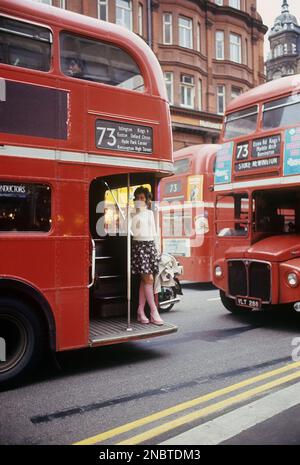 London 1972. Blick auf die Oxford Street mit den typischen roten Doppeldeckerbussen. Eine junge Frau wird auf dem Bahnsteig gesehen, bereit zum Abheben. Sie trägt ein typisches 1970er-Outfit, einen Pullover, einen kurzen rosa gemusterten Rock und dazu passende pinkfarbene Lederstiefel in einem Plattformmodell. Eine Schuhmode, die zu dieser Zeit in Europa und Großbritannien beliebt war und bis 1976, als Plateauschuhe plötzlich aus der Mode kamen, so blieb. Dieses Jahr trugen 1972 berühmte Leute wie David Bowie, David Johansen von den New York Dolls Plateauschuhe. Kristoffersson Ref. DV2 Stockfoto