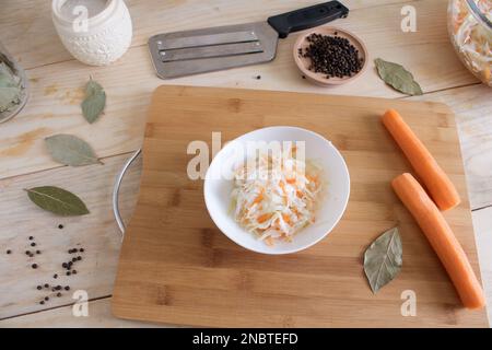 Heim-Sauerkraut mit Karotten und Gewürzen auf einem Teller, Sauerkraut. Gemischter Salat aus frischem Gemüse auf einem Teller mit einer Gabel auf Holzhintergrund. Stockfoto