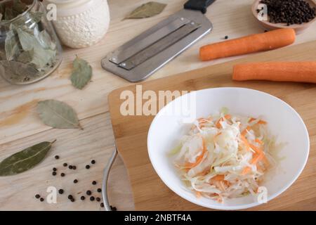 Heim-Sauerkraut mit Karotten und Gewürzen auf einem Teller, Sauerkraut. Gemischter Salat aus frischem Gemüse auf einem Teller mit einer Gabel auf Holzhintergrund. Stockfoto